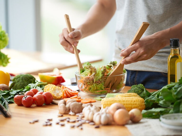 A man tosses a healthy salad, healthy food spread across the kitchen counter like tomatoes, brocoli, spinach, carrots, and more. GLP-1 near Colleyville, TX, should be paired with healthy eating habits like this to ensure healthy weightloss. 